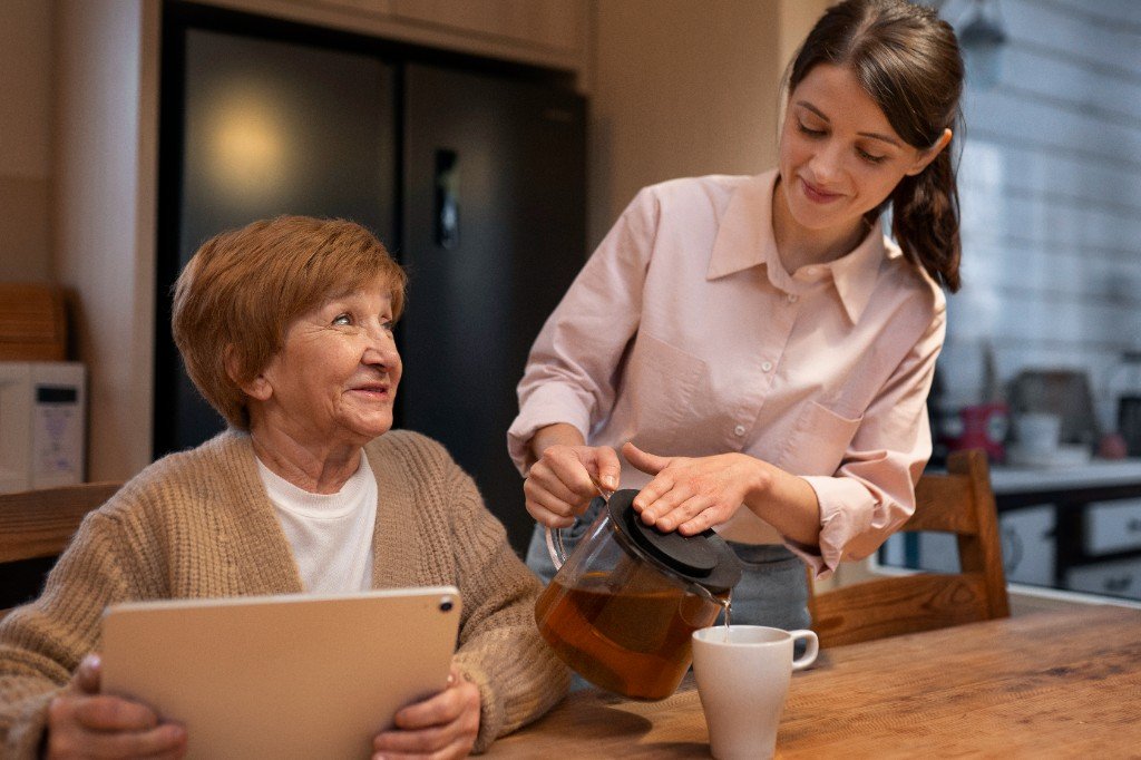 Care worker pouring a drink for an elderly woman at home, sharing a warm moment of companionship